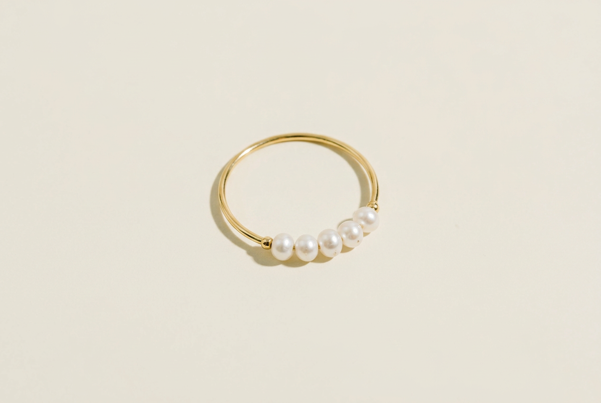 Freshwater Pearl Ring, thin gold band ring with a row of small white freshwater pearls on a warm cream studio background